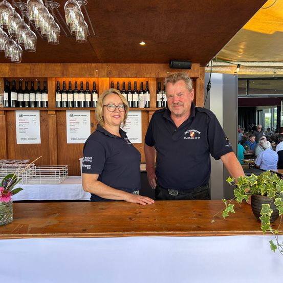 Eine lächelnde Frau und ein Mann, beide in blauen Uniformen, stehen hinter einer Bartheke in einem Restaurant. Hinter ihnen befinden sich Regale mit Weinflaschen und im Hintergrund sitzende Gäste an Tischen.