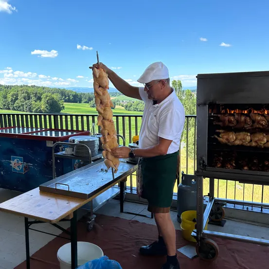Ein Mann grillt auf einem Balkon. Er hält einen Spieß mit Hühnchen und trägt einen weißen Hut und grüne Shorts. Ein Grill mit mehr Hühnchen steht in der Nähe. Bäume und ein Feld sind im Hintergrund.
