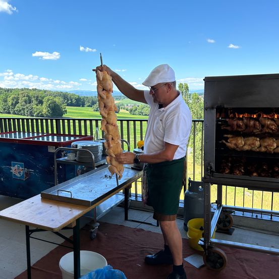 Ein Mann grillt auf einem Balkon. Er hält einen Spieß mit Hühnchen und trägt einen weißen Hut und grüne Shorts. Ein Grill mit mehr Hühnchen steht in der Nähe. Bäume und ein Feld sind im Hintergrund.