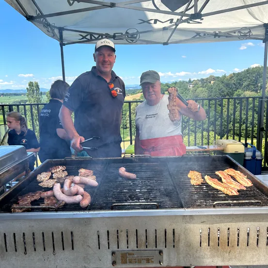 Zwei Männer grillen Würste und Fleisch auf einem Outdoor-Grill, mit Menschen und Bäumen im Hintergrund. Ein Mann hält eine Wurst mit Zange.