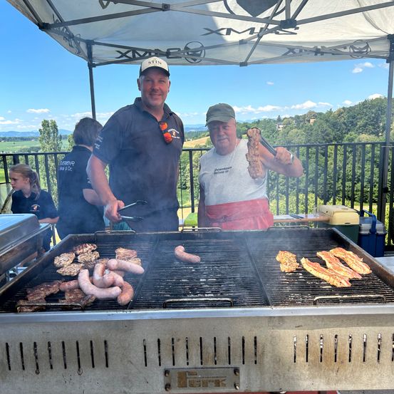 Zwei Männer grillen Würste und Fleisch auf einem Outdoor-Grill, mit Menschen und Bäumen im Hintergrund. Ein Mann hält eine Wurst mit Zange.
