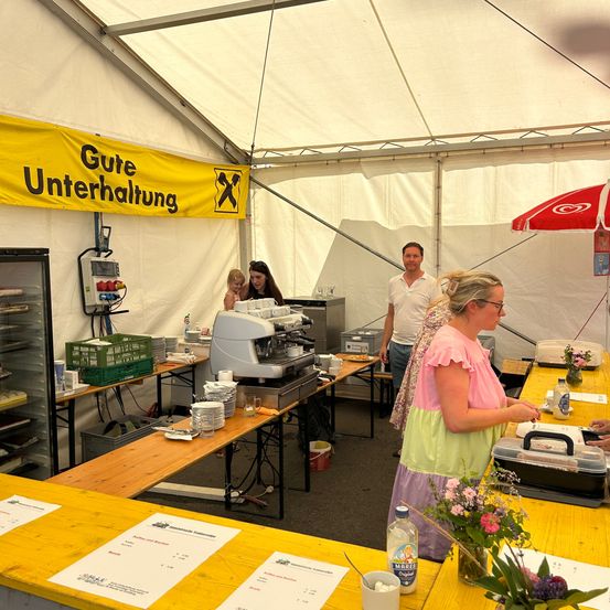 Ein Essensstand unter einem Zelt mit einem gelben Banner, das 'Gute Unterhaltung' steht. Im Inneren steht eine Frau an einem Tresen mit Blumen, während ein Mann zuschaut. Ein Kind und eine weitere Frau sind in der Nähe. Tische sind mit Tellern und einer Kaffeemaschine besetzt.