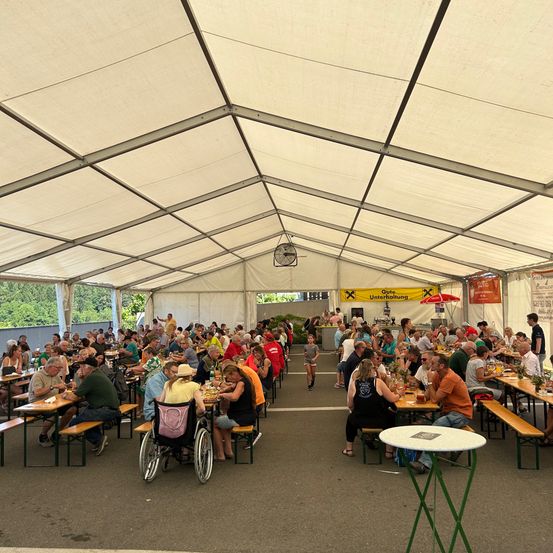 Ein großes Zelt mit vielen Menschen, die an Tischen sitzen und essen, mit einem kleinen Kind, das zwischen ihnen hindurchläuft. Das Zelt hat einen Metallrahmen und ist mit einem weißen Stoff bedeckt.