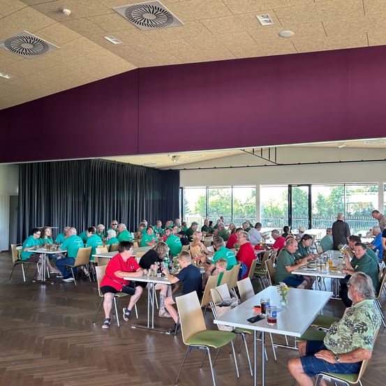 Viele Menschen sitzen an Tischen in einem Essbereich, tragen grüne und rote Hemden. Der Raum hat einen Holzboden, Glasfenster und eine purpurfarbene Wand.