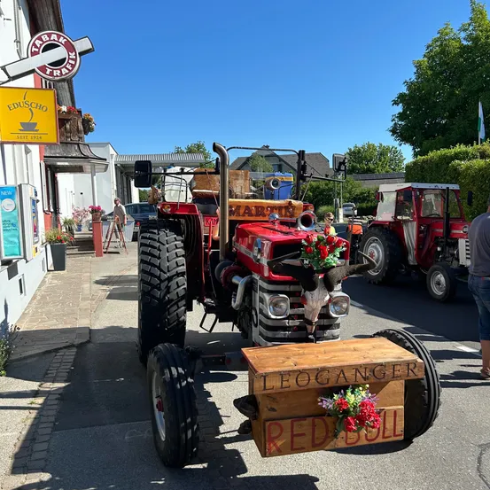 Ein dekorierter roter Traktor mit Blumen und einem Kuhschädel auf einem Anhänger steht Leoganger Red Bull. Er ist auf der Straße geparkt, mit einem weiteren Traktor dahinter.