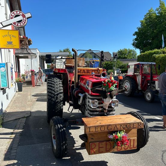 Ein dekorierter roter Traktor mit Blumen und einem Kuhschädel auf einem Anhänger steht Leoganger Red Bull. Er ist auf der Straße geparkt, mit einem weiteren Traktor dahinter.