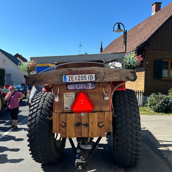 Die Rückseite eines Traktors mit dem Kennzeichen ZE 205 ID, auf einer Straße geparkt. Dahinter steht eine Frau, und verschiedene Gebäude sind sichtbar.