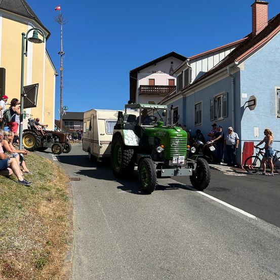 Ein grüner Traktor fährt mit einem Wohnwagen hinter sich die Straße hinunter. Zuschauer beobachten von der Seite. Eine Flagge ist an einem Pfahl in der Nähe.