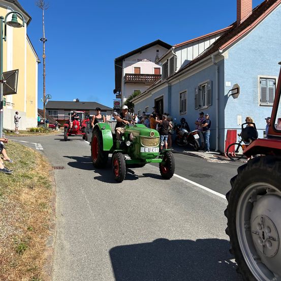 Ein grüner Traktor fährt die Straße entlang, während Menschen vom Bürgersteig zusehen. Dahinter folgt ein weiterer Traktor. Gebäude säumen die Straße, und ein Fahrrad ist in der Nähe geparkt.