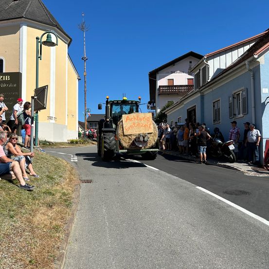 Ein Traktor fährt mit einem Heuballen an der Front durch eine Dorfstraße. Zuschauer sind an den Straßenrändern versammelt. Gebäude mit Balkonen und Schildern sind sichtbar.