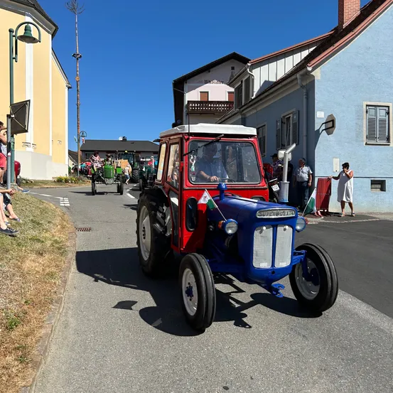 Ein blauer und roter Traktor fährt eine Straße hinunter, gesäumt von Gebäuden und Menschen. Eine Person im Traktor winkt mit Flaggen. Zuschauer stehen am Bürgersteig, und eine Frau steht in der Nähe eines blauen Gebäudes.