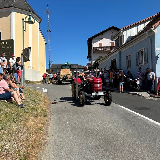 Ein roter Traktor fährt auf der Straße in einem Dorf. Die Leute schauen an den Seiten zu. Ein Schild mit dem Text 'ZIB Nikolai' steht auf der linken Seite.