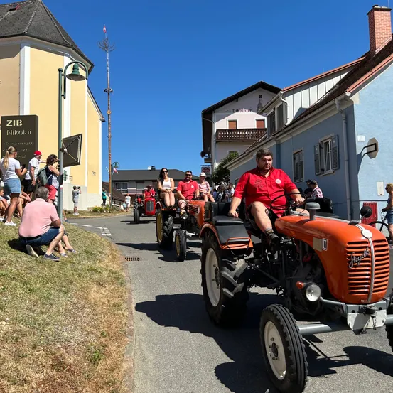 Eine Traktorparade mit mehreren roten Traktoren auf einer Straße, Zuschauer schauen zu. Gebäude und ein klarer Himmel im Hintergrund.