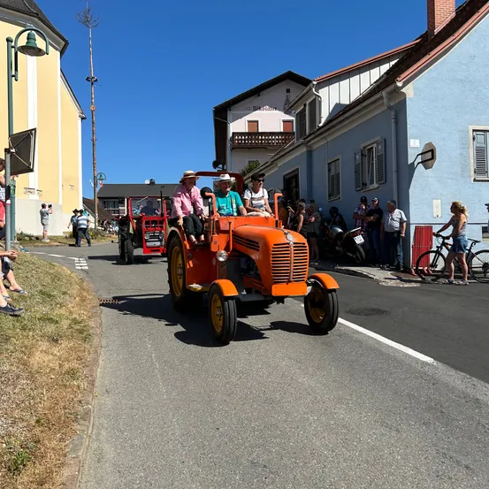 Zwei Personen fahren auf einem orangefarbenen Traktor auf einer Straße mit Zuschauern, Fahrrädern und Gebäuden.