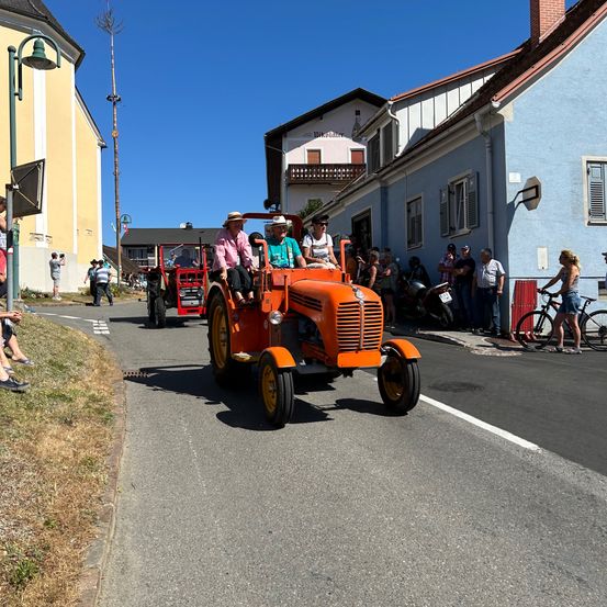 Zwei Personen fahren auf einem orangefarbenen Traktor auf einer Straße mit Zuschauern, Fahrrädern und Gebäuden.