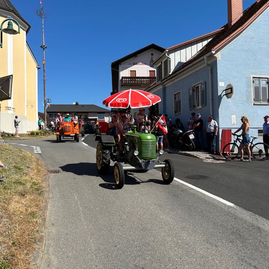 Eine Gruppe von Menschen fährt auf einem grünen Traktor mit einem roten Regenschirm an einem sonnigen Tag. Leute beobachten am Straßenrand.