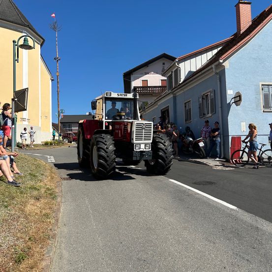 Ein Traktor fährt auf einer Straße in einer Kleinstadt, mit Zuschauern am Bürgersteig und Gebäuden auf beiden Seiten. Der Himmel ist klar und blau.