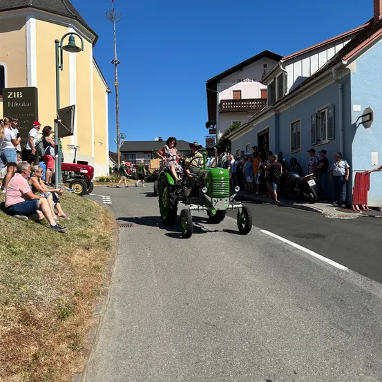 Ein grüner Vintage-Traktor fährt auf einer Straße in einer Kleinstadt, mit Zuschauern an den Seiten. Ein Gebäude mit einem Schild, das ZIB Nikolau liest, ist in der Nähe.