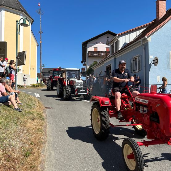 Ein Mann fährt einen roten Traktor in einer Parade. Zuschauer säumen die Straße. Ein Gebäude mit einer Flagge und einem hohen Mast befindet sich in der Nähe. Ein weiterer Traktor folgt dahinter.
