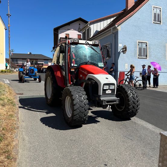 Ein roter Traktor fährt vor einem blauen Haus die Straße hinunter. Mehrere Menschen stehen am Bürgersteig. Ein Fahrrad ist in der Nähe geparkt.