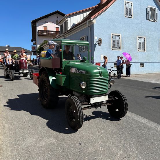 Ein grüner Traktor fährt auf einer Straße in einer Stadt. Mehrere Personen fahren darauf, und andere beobachten von der Straßenseite. Ein blaues Haus mit Fensterläden steht hinter dem Traktor.