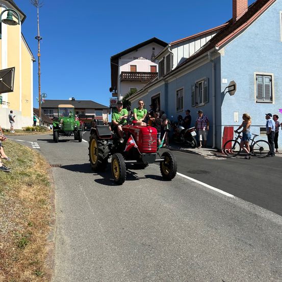 Ein roter Traktor mit zwei Personen in grünen T-Shirts fährt die Straße hinunter, gefolgt von einem grünen Traktor. Zuschauer säumen die Straße und beobachten den Umzug.