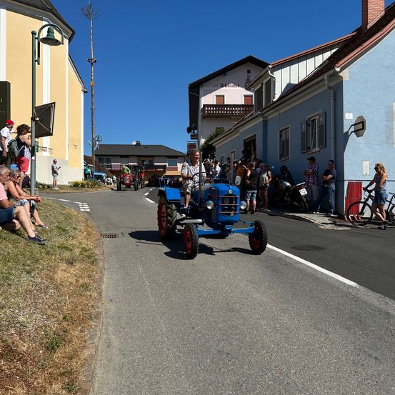 Ein blauer Traktor fährt eine Straße hinunter, mit einem Mann am Steuer, während Zuschauer an der Seite stehen. Eine Menge beobachtet vom Gras aus, Gebäude und geparkte Fahrzeuge säumen die Straße.