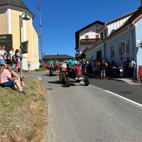 Eine Gruppe von Menschen beobachtet einen Oldtimer-Traktoren-Parade an einem sonnigen Tag, mit Zuschauern, die am Straßenrand sitzen.