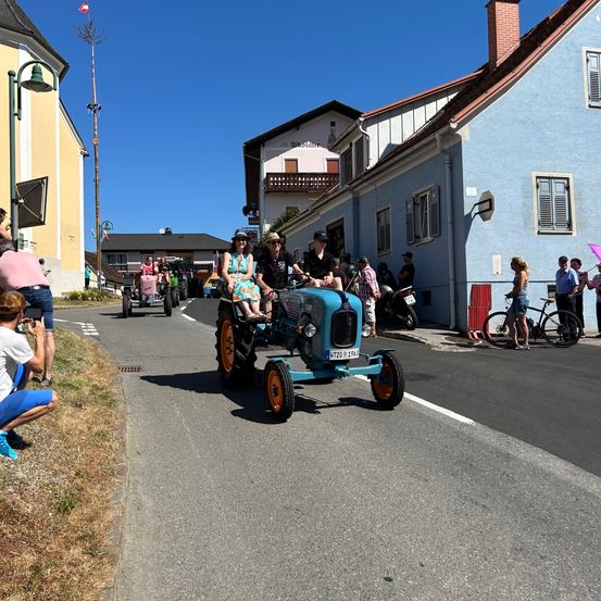 Ein alter blauer Traktor fährt eine Straße hinunter, mit Zuschauern auf beiden Seiten. In der Nähe ist ein weiterer Traktor zu sehen, auf dem Menschen sitzen. Gebäude säumen die Straße, und einige Zuschauer sitzen oder stehen auf dem Bordstein.