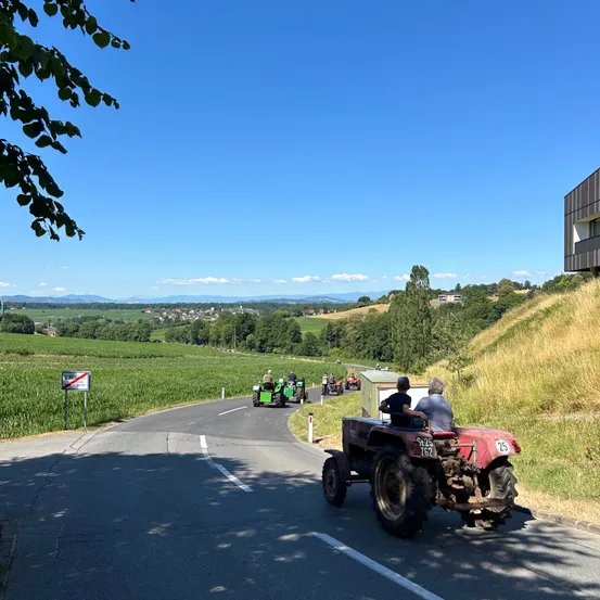 Mehrere Oldtimer-Traktoren fahren auf einer Landstraße mit Blick auf grüne Felder, Berge und ein modernes Gebäude im Hintergrund.