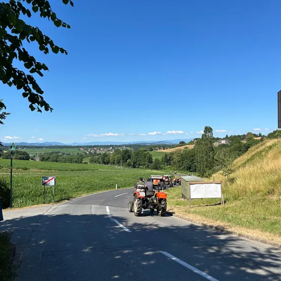 An einem sonnigen Tag fährt eine Gruppe von Menschen Traktoren auf einer ländlichen Straße, umgeben von einem grünen Feld und entfernten Bergen.