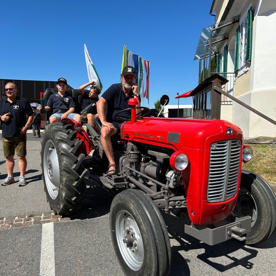 Eine Gruppe von Menschen fährt draußen auf einem roten Traktor, wobei ein Mann fährt. In der Nähe ist ein Gebäude mit Fahnen zu sehen, und ein Mann steht an der Seite.