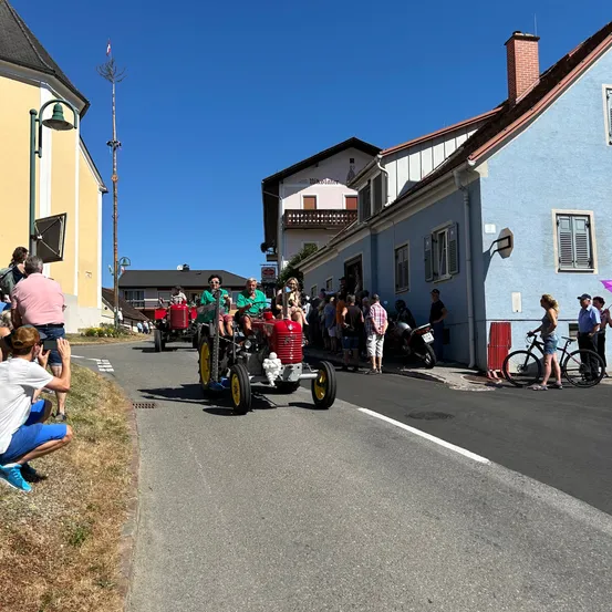 Menschen beobachten, wie eine Parade roter Traktoren an einem sonnigen Tag vorbeizieht. Zuschauer säumen die Straße, einige auf Fahrrädern. Die Traktoren werden von Personen in grünen Hemden gefahren.