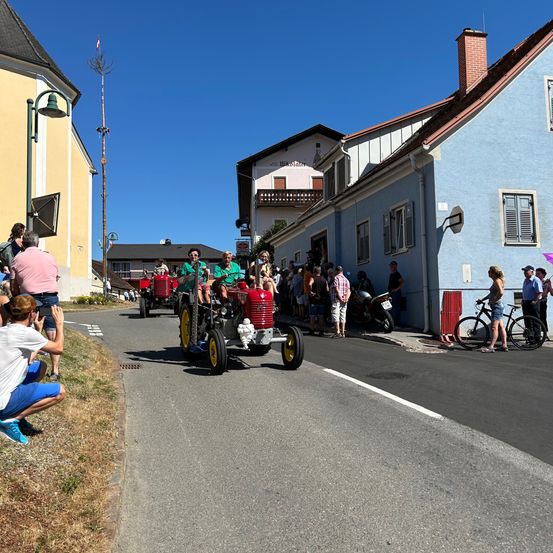 Menschen beobachten, wie eine Parade roter Traktoren an einem sonnigen Tag vorbeizieht. Zuschauer säumen die Straße, einige auf Fahrrädern. Die Traktoren werden von Personen in grünen Hemden gefahren.