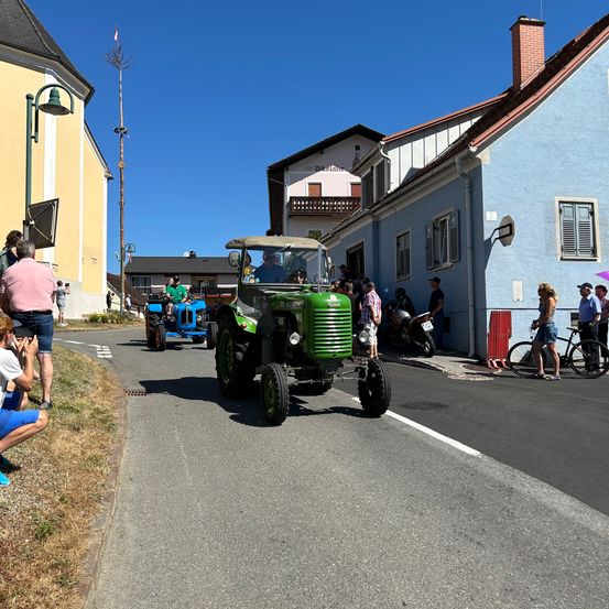 Eine Parade von Oldtimer-Traktoren auf einer Straße mit Zuschauern an der Seite. Eine Person hockt und eine andere hält eine Kamera. Ein blauer Traktor folgt einem grünen.