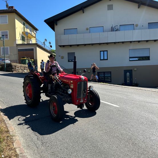Ein Mann fährt einen roten Traktor die Straße hinunter, während eine Frau ein Foto am Bürgersteig macht. Im Hintergrund sind Gebäude und geparkte Autos zu sehen.