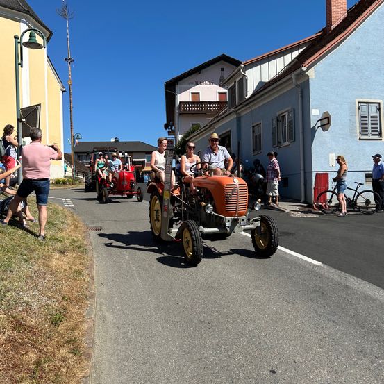 Menschen fahren mit Traktoren auf der Straße. Zwei Traktoren fahren die Straße entlang. Zuschauer stehen an der Seite.