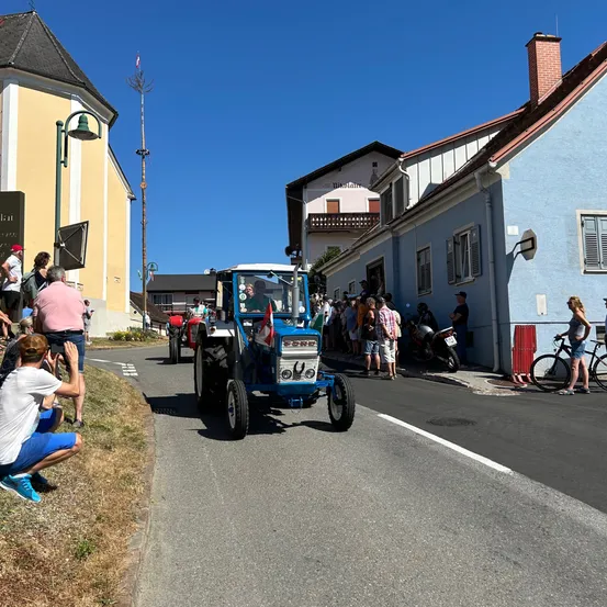 Ein blauer Traktor fährt eine Straße hinunter, an der eine Menschenmenge auf dem Bürgersteig steht. Ein Mann macht ein Foto und ein anderer fährt ein Fahrrad. Die Gebäude haben Balkone und Schornsteine.