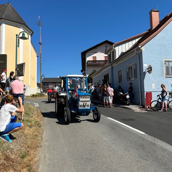 Ein blauer Traktor fährt eine Straße hinunter, an der eine Menschenmenge auf dem Bürgersteig steht. Ein Mann macht ein Foto und ein anderer fährt ein Fahrrad. Die Gebäude haben Balkone und Schornsteine.