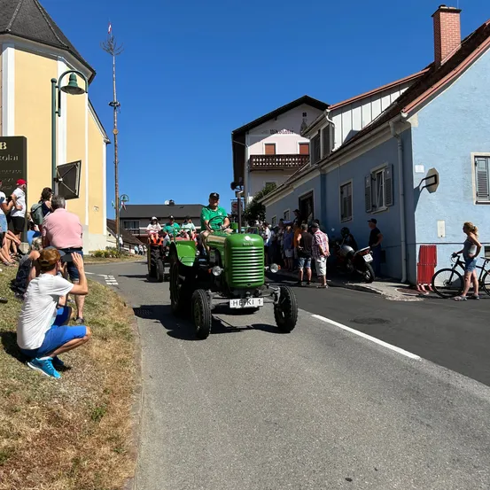 Ein grüner Traktor fährt auf einer Straße, die von mehreren Menschen beobachtet wird. Einige stehen auf dem Bürgersteig, andere auf dem Rasen. Der Traktor hat ein Nummernschild mit der Aufschrift H5K1.