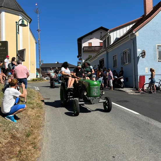 Ein grüner Oldtimer-Traktor fährt eine Straße hinunter, gesäumt von Zuschauern, Gebäuden und geparkten Fahrrädern. Die Leute stehen am Straßenrand, einige machen Fotos. Dem Traktor folgt ein weiterer.