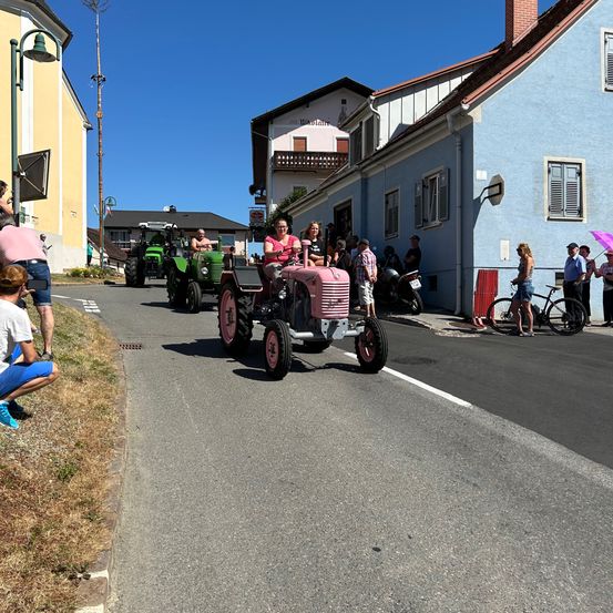Einige Traktoren fahren mit Zuschauern an der Seite die Straße hinunter. Eine Frau fährt einen rosa Traktor.