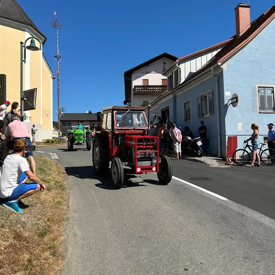 Ein roter Traktor fährt die Straße hinunter, während viele Menschen zusehen. Ein Mann kniet. Ein Motorrad ist vor einem Gebäude geparkt.