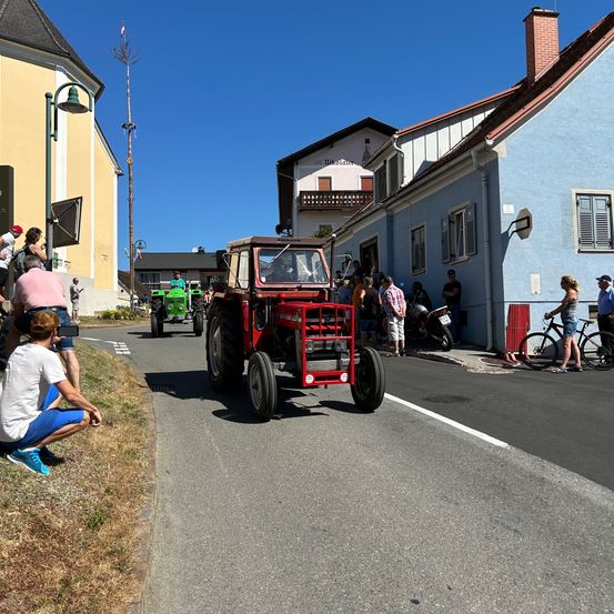 Ein roter Traktor fährt die Straße hinunter, während viele Menschen zusehen. Ein Mann kniet. Ein Motorrad ist vor einem Gebäude geparkt.