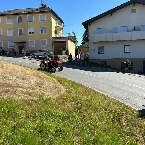 Ein kleiner roter Traktor fährt auf einer Straße, an der Menschen auf dem Bürgersteig stehen. Es gibt zwei Gebäude, eines gelb und eines weiß, am Straßenrand.