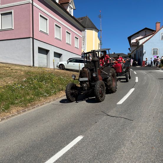 Zwei alte Traktoren fahren eine Straße hinunter, mit Gebäuden auf beiden Seiten. Zuschauer säumen die Straße und beobachten, wie ein weißes Auto vorbeifährt.