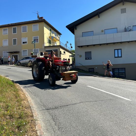 Ein roter Traktor fährt vor zwei Gebäuden mit Menschen, die draußen stehen und sitzen.