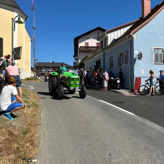 Ein grüner Traktor fährt auf einer Straße in einem Dorf, mit Zuschauern an den Seiten, einer Person, die kniet, und einer anderen mit einem Fahrrad.