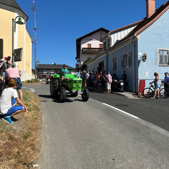 Ein grüner Traktor fährt auf einer Straße in einem Dorf, mit Zuschauern an den Seiten, einer Person, die kniet, und einer anderen mit einem Fahrrad.