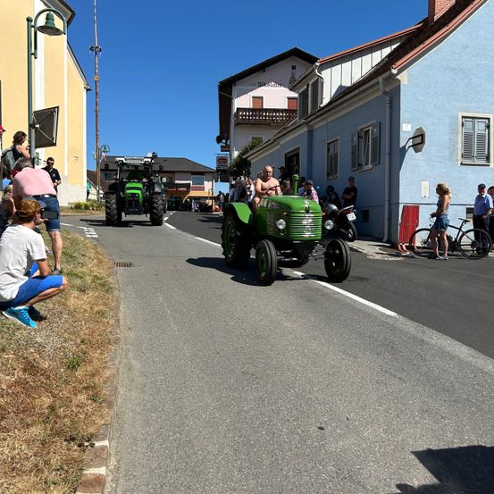 Ein grüner Traktor fährt auf einer Straße in einer Kleinstadt vorbei, während Zuschauer am Straßenrand stehen. Im Hintergrund befinden sich Gebäude und ein Fahnenmast.